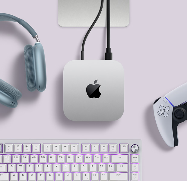Pohled shora na stříbrný Mac mini se dvěma kabely připojenými k portům na zadní straně, vedle jsou AirPody Max, stříbrný Magic Keyboard a bezdrátový herní ovladač